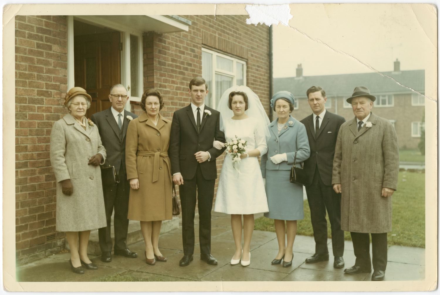 An aged colour photo of a wedding party of eight standing outside a suburban house in 1966, faded and warm-shifted, with a crease and a patch of emulsion loss at the top corner.
