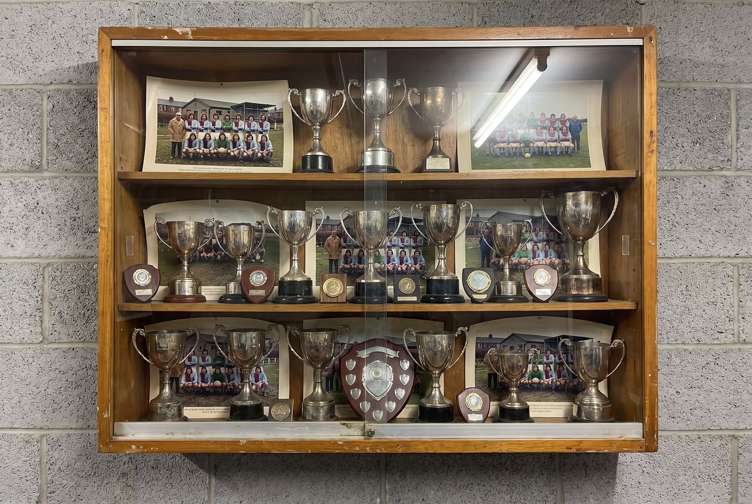 A glazed wooden trophy cabinet on a clubhouse wall, three shelves of small tarnished cups and shields, with curled framed team photos propped behind them.