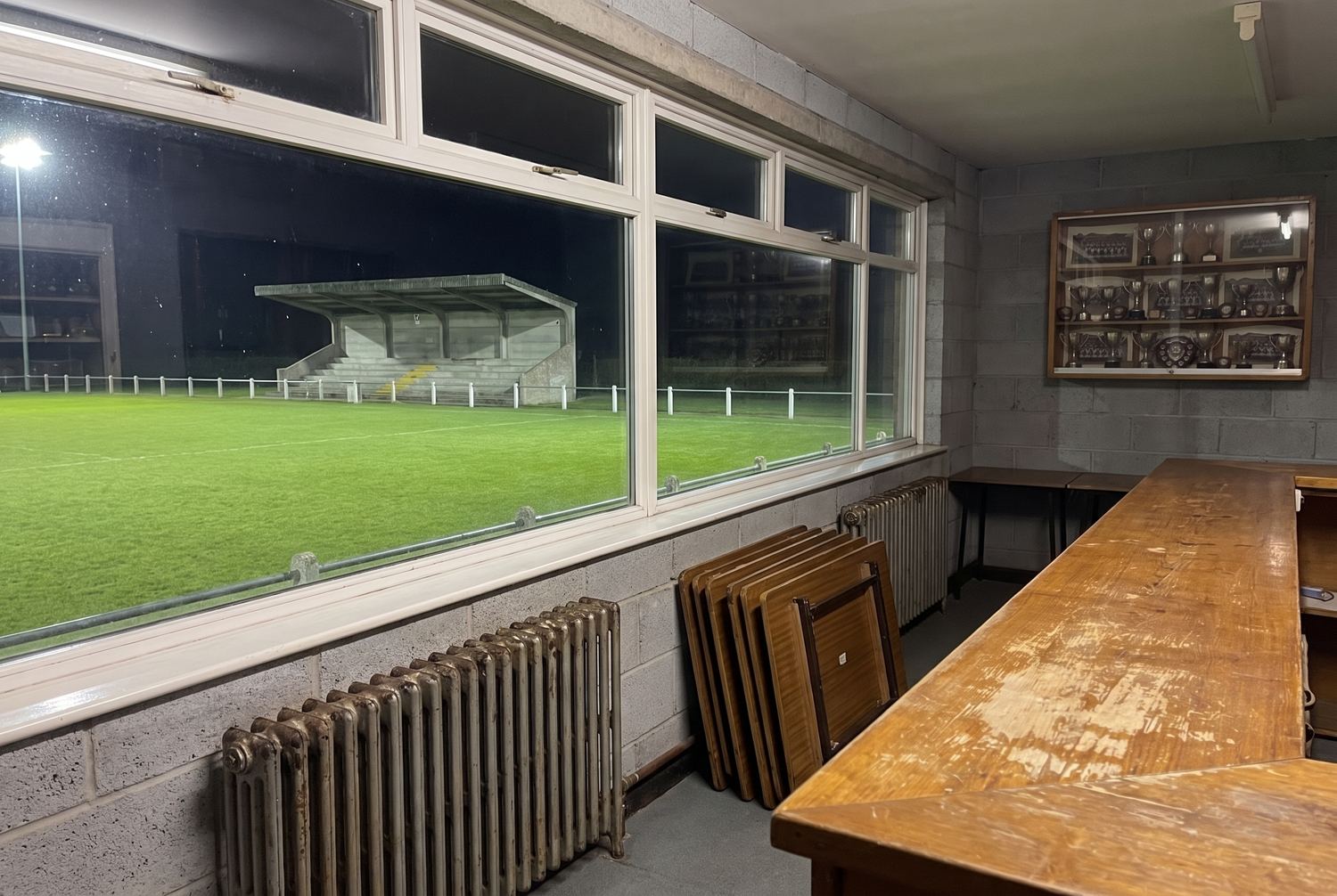 The inside of a small football clubhouse, looking out through a long window at a floodlit pitch and a small covered stand.