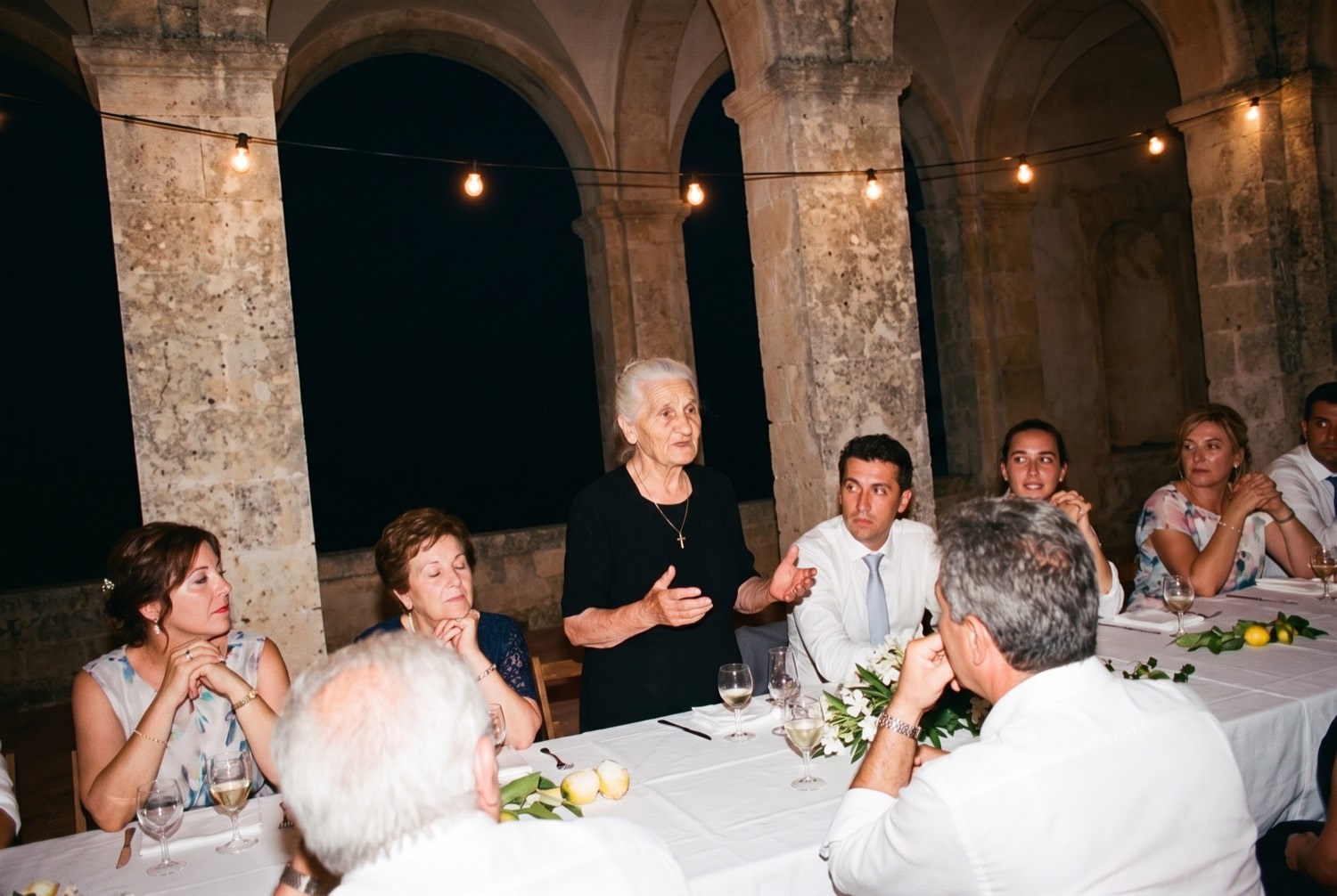 A white-haired woman in black standing to speak at a long table at night, faces lit hard by a camera flash.