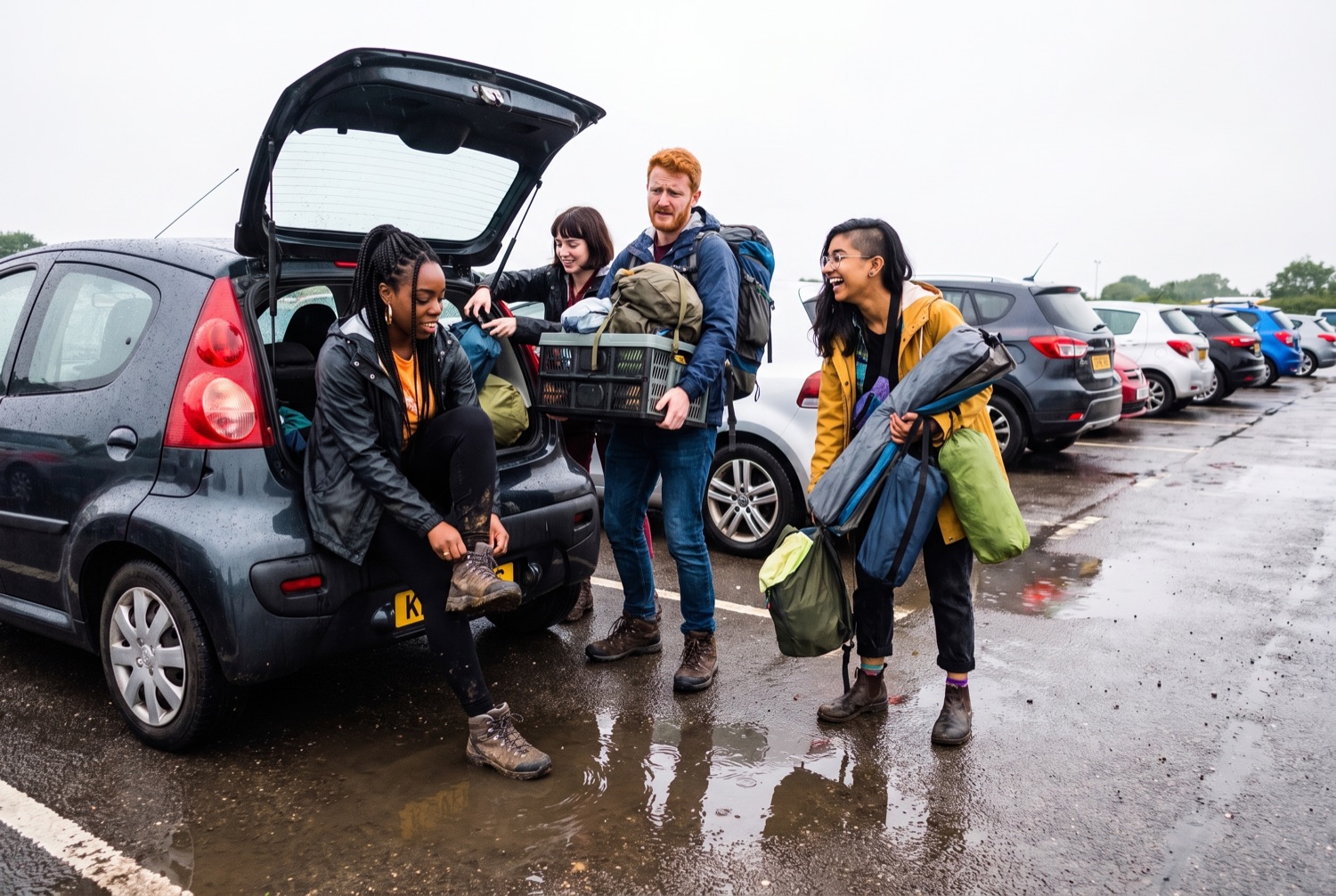 Four friends unloading a small hatchback in a wet car park, pulling out rucksacks, a folded tent and a crate.