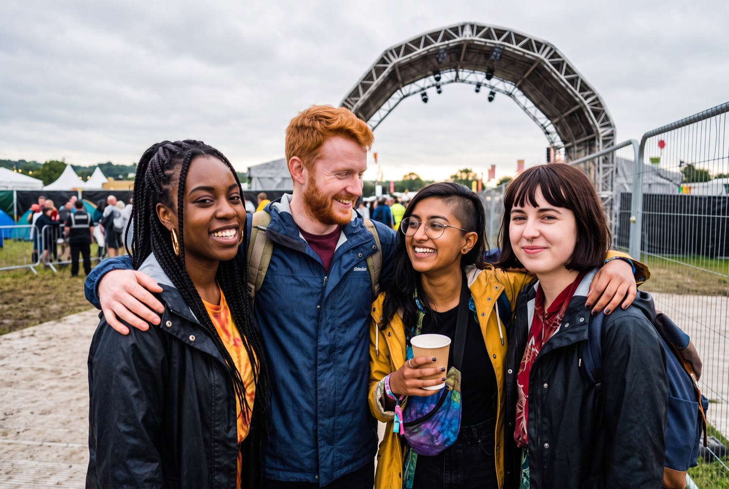 Four friends in their late twenties standing arm in arm in front of a festival entrance arch, bags still on their backs.