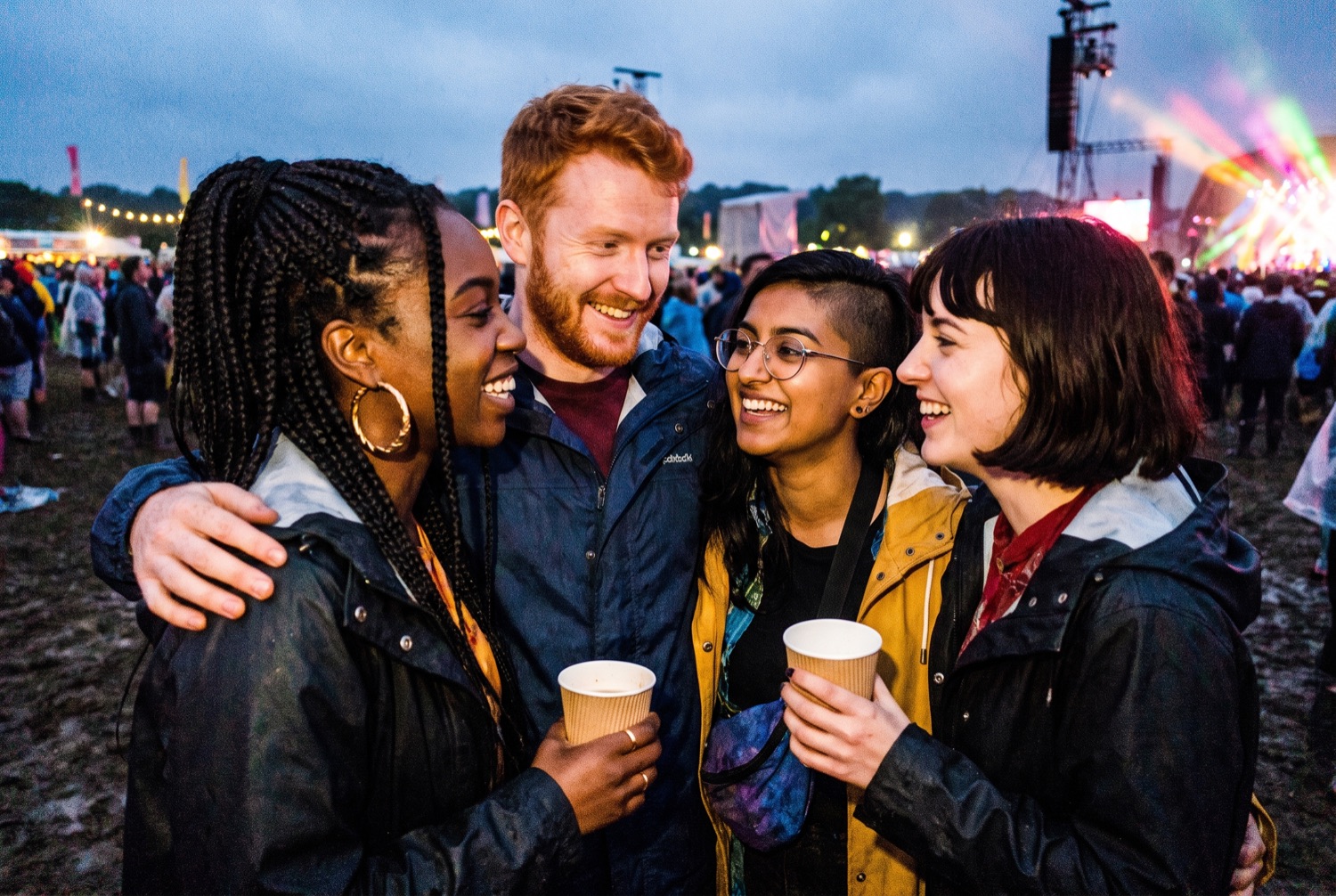 Four friends standing arm in arm in a festival crowd at dusk, two of them holding paper cups, a lit stage behind them.