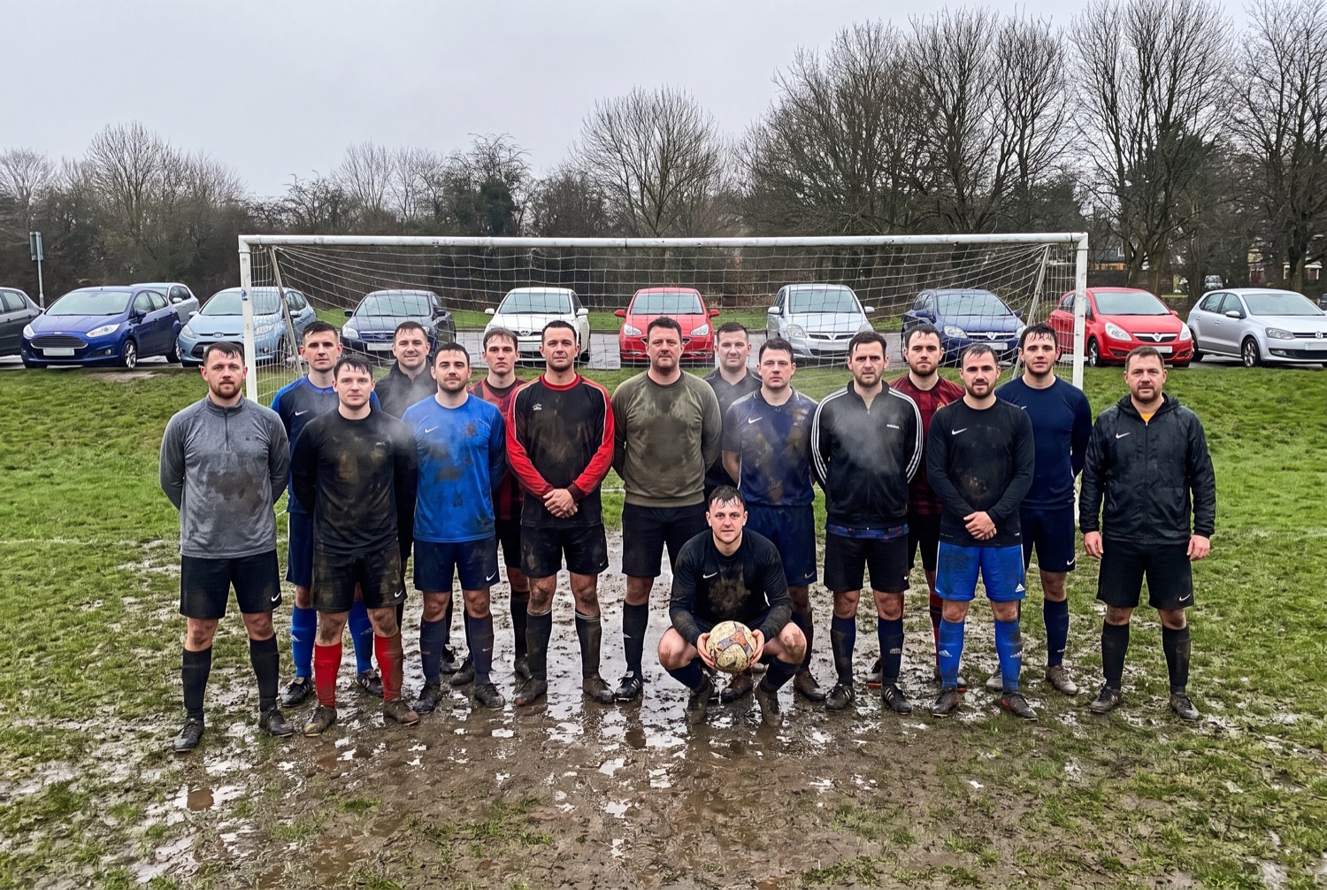 An amateur eleven-a-side team in mismatched training tops standing in two rows on a wet municipal pitch, breath visible in the cold.